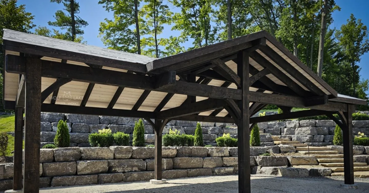 Covered timber structure built with dark wood, standing on a gravel base in a landscaped backyard.